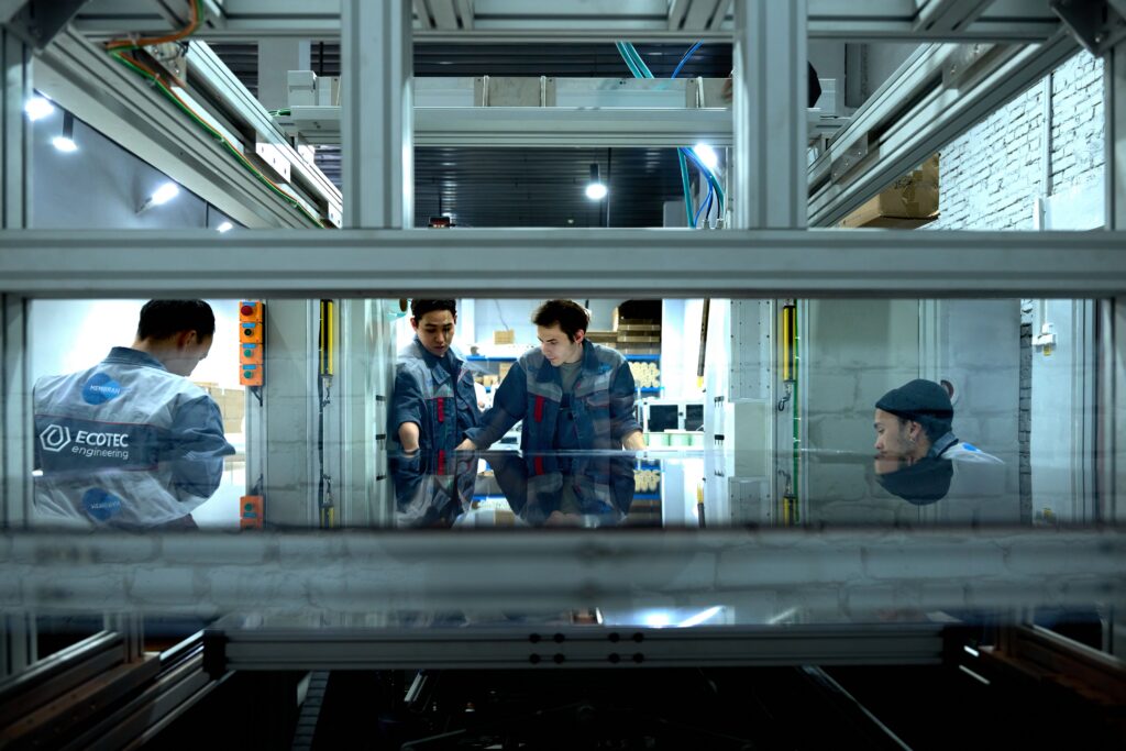 Industrial workers in matching uniforms examining a large glass panel inside a manufacturing facility frame